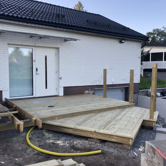 Nybyggd frukostaltan i trä med unika vinklar, skrapgaller och infälld spolbrunn framför vit södervänd husfasad.