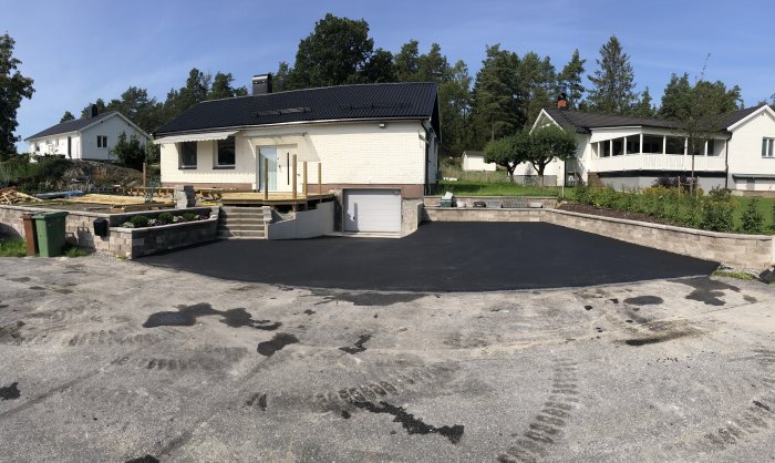 Ny asfalterad uppfart framför vit husfasad med garage och trädgård i bakgrunden.