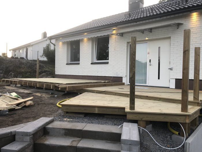 Pågående arbete med trädäck längs en vit tegelvilla, konstruktion och trävirke synligt.