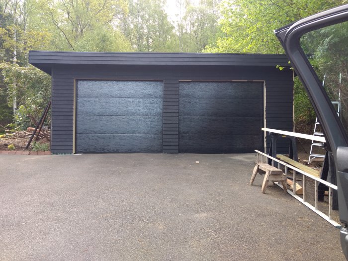 En nästan färdigställd svart garagebyggnad med en monterad port och arbetsmaterial bredvid.
