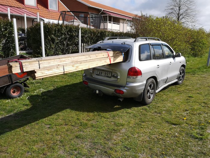 Bil parkerad med släpvagn lastad med långa träläkt som sticker ut bakifrån bilen.