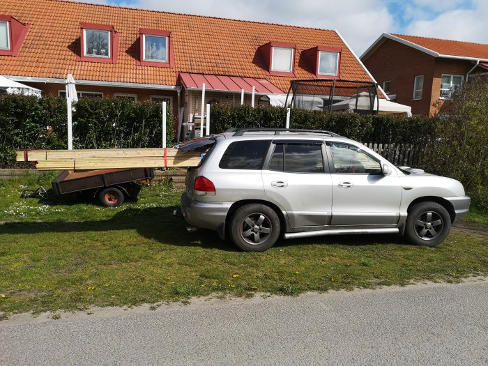 Bil med släp lastat med långa träreglar, parkerad vid hus och trädgård.