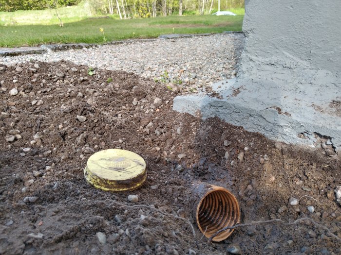 Dräneringsrör i jord med ett gult lock från Plastic Padding vid husgrund.