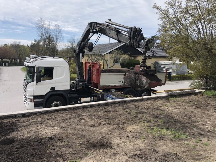 Lastbil med kran som lossar jord på en tomt för gräsmatteanläggning.