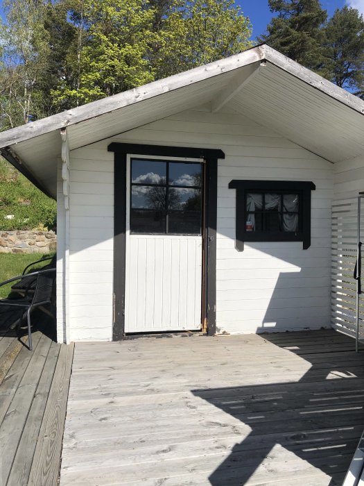 Vitmålad förrådsbyggnad med en nyinstallerad dörr och ett fönster, beläget i en trädgård.