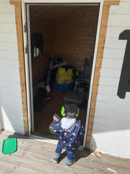 Ett barn står framför en öppen förrådsdörr som leder in till ett rörigt förråd, i dagsljus.