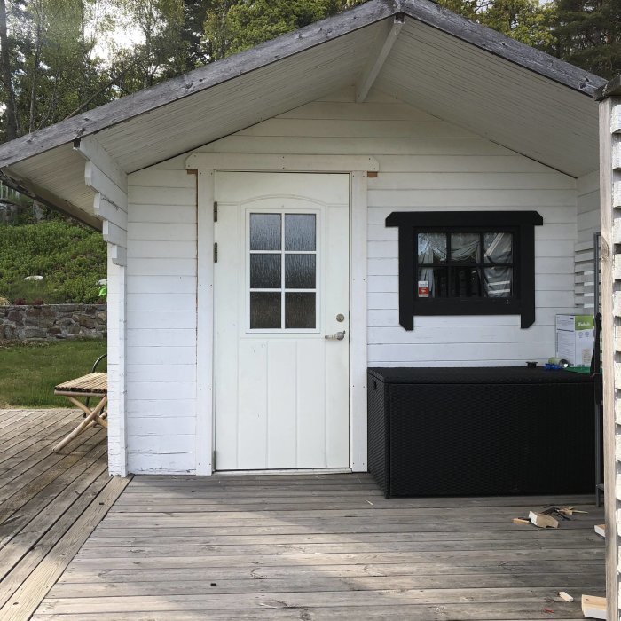 Renoverat vitmålat förråd med ny dörr och svart kantat fönster, veranda framför.