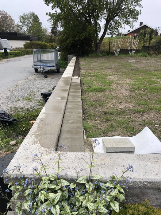 Ouppbyggd stenkant längs en gräsmatta med en vagn och blommande växter i förgrunden, under pågående arbete.