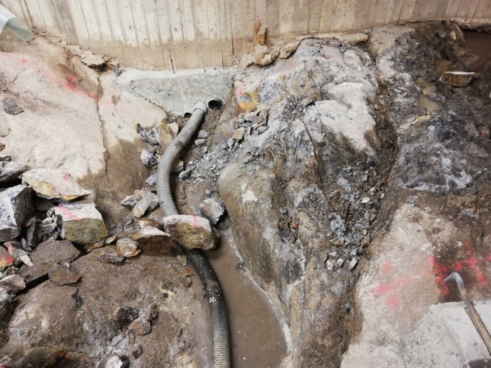 Källarutgrävning med stora stenblock och dräneringsslang mot betongvägg.