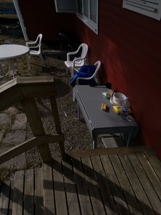 Trädäck med utemöbler och grill intill en röd vägg i trädgården, soligt väder.