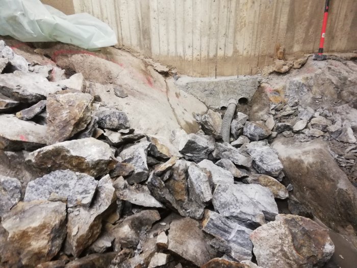 Stenhög och krossade stenblock vid grävarbete nära betongvägg och rörledning.