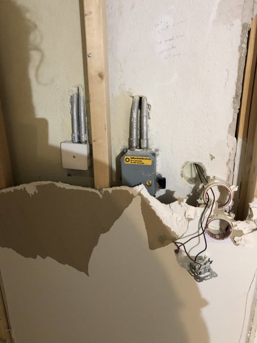 Vägg i källare med synlig skada och fukt, exponerade elledningar och ventilationsrör, samt nerskrivna noteringar.