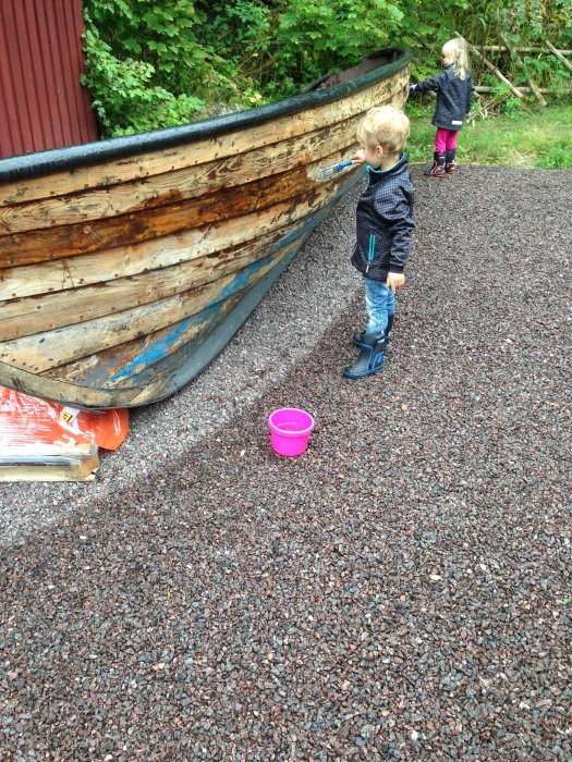 Två barn leker vid en gammal träbåt som står omvandlad till lekstuga på grusmark.