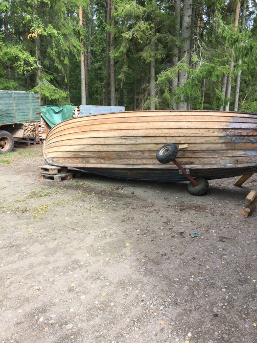 Omvänd träbåt som står på pallar och väntar på nedslipning, omgiven av skog och en trailer i bakgrunden.