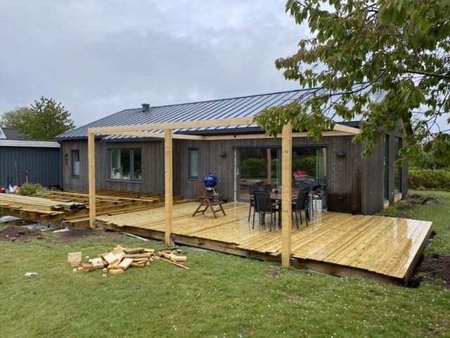 Pågående konstruktion av en träaltan med en pergola utan tak bredvid huset, synliga reglar och några stolpar, trädgård i bakgrunden.