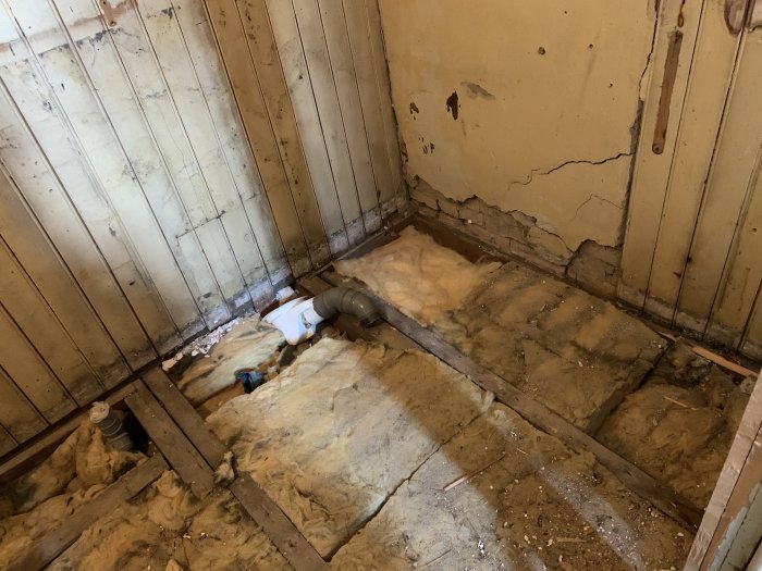 Rivet badrumsgolv med blottade träbjälkar, isolering och rörledningar före renovering.