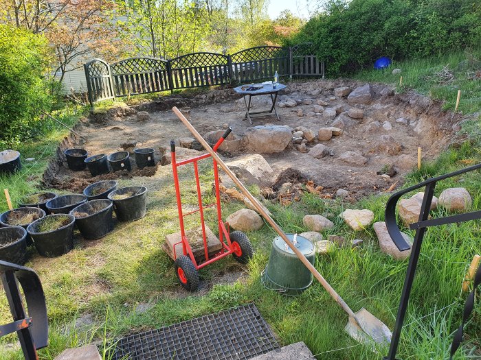 Trädgård under ombyggnad med spräckta stenblock, trädgårdsverktyg och hinkar med jord.