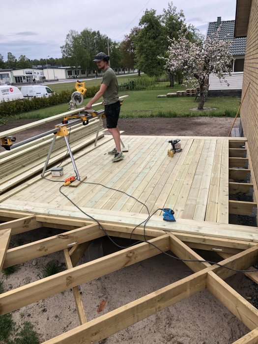 Person bygger träterrass med kapmaskin och verktyg ute i trädgård.