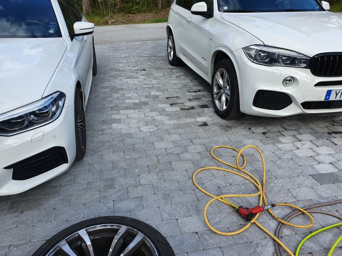 Två bilar parkerade på stenläggning bredvid lös sommardäck och gul förlängningssladd.