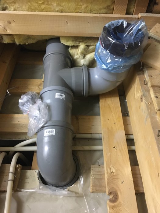 Nytt avloppsrör installerat mellan träbjälkar med stödregling och isolering i bakgrunden.