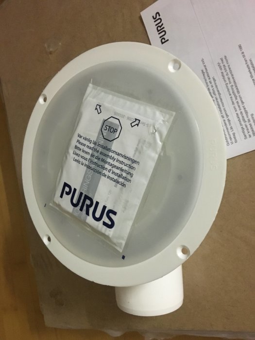 En Purus golvbrunn i plast med monteringsinstruktioner, paketerad och placerad på ett brunt underlag.