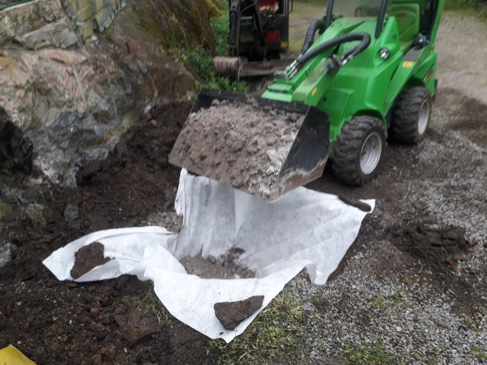 Grävmaskin tömmer grus på markduk vid stenplintar med armeringskorgar i en grävd grop.