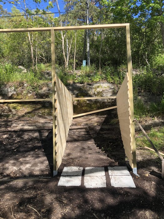 Påbörjat träregelverk för ett byggprojekt står på betongplattor utomhus med skog i bakgrunden.