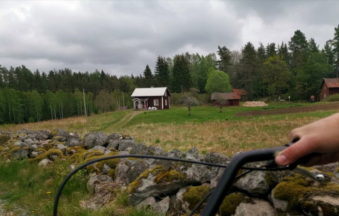 Vy från en moped av en lantlig skogsstig och röd stuga med vita knutar, omgiven av träd och stenmur.