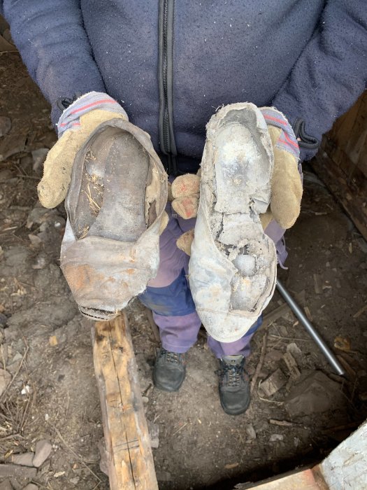 Person håller i tva gamla, slitet par ballerinaskor mot en bakgrund av murket timmer och grus.