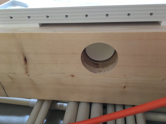 Ett 55mm borrat hål i ett träblock, ovanpå vita rörledningar, för montering av Purus avloppsbrunn.