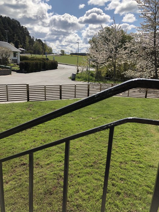 Utsikt från en veranda över en välskött gräsmatta med blommande träd och en klarblå himmel med fluffiga moln i bakgrunden.