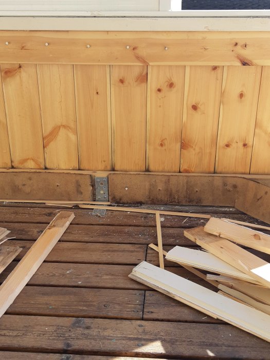 Delvis demonterad träpanel på en veranda med synliga skruvar och några ruttna brädor på golvet.