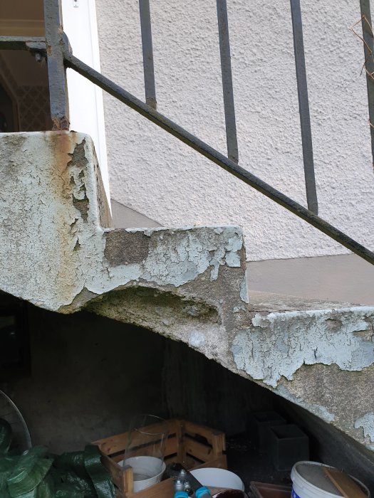 Trappe med afskallet beton og rustne gelændere, viser skader under trappen. Trærammer og spande ses i baggrunden.