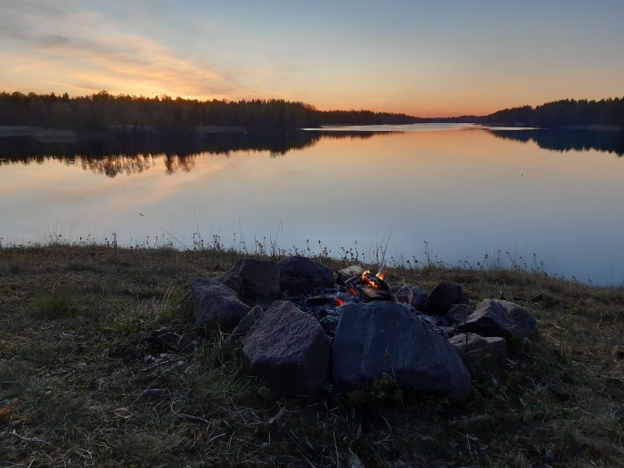Solnedgång över Närdingen med en slocknande brasa i förgrunden och spegelblank sjö i Roslagen.