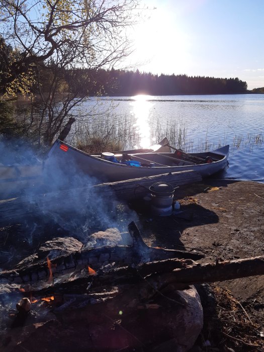 En kanot vid sjökanten nära en brinnande lägereld under solnedgången vid Aspdalssjön.