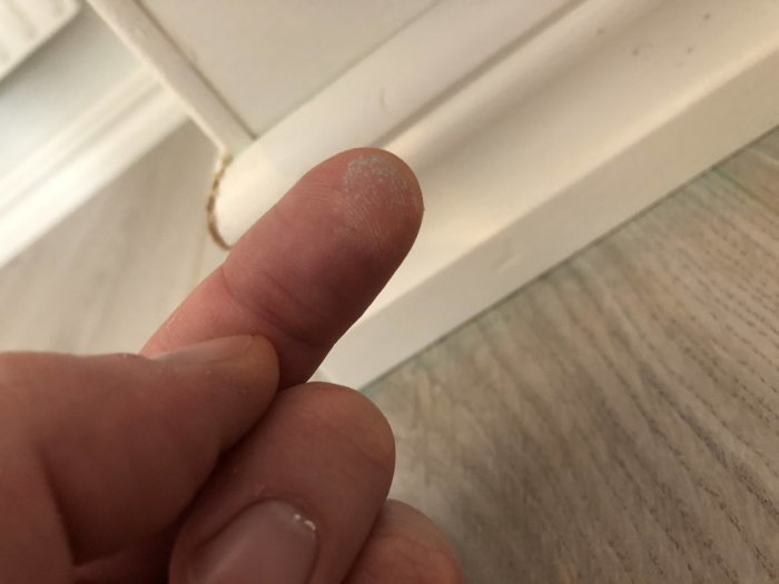 Grönt pulver på ett finger mot bakgrund av ett laminatgolv och vit golvsockel.