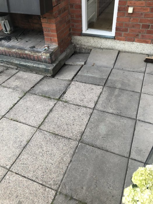 Ojämna betongplattor på uteplats nära husvägg och trappsteg, i behov av justering.