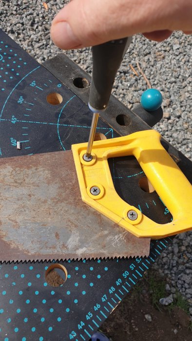 Hand som använder en skruvmejsel för att demontera ett gult handtag på en rostig gammal såg som ligger på ett bord med gradmätning.