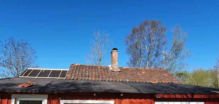 Hus med tegeltak och synliga svackor, skorstenspipa, solpaneler och omgivande träd mot klarblå himmel.