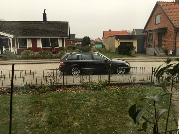 BMW E61 525i parkerad utanför ett hus, sett från ett fönster över en gräsmatta och staket.