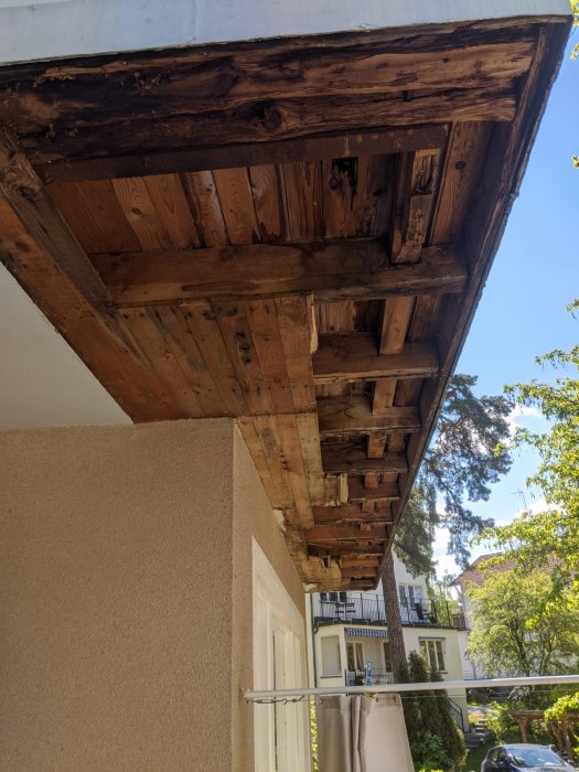 Undersidan av en gammal skadad balkong med träbjälkar och råspont, vattenskador syns nära hängränna.
