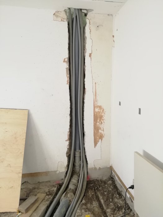 Vertikalt rörschakt med flera elrör i hörn av rum nära oskyddad betongvägg och spånplatta.
