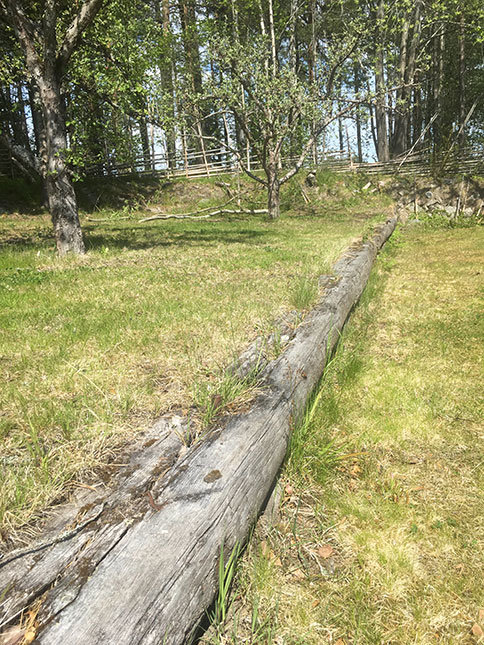 Gammal träsliper mur som sträcker sig genom en solig gräsmatta med träd i bakgrunden.