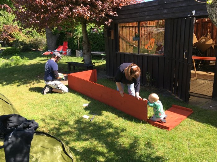 Familj engagerad i trädgårdsarbete, målar röda odlingslådor ute i solskenet.