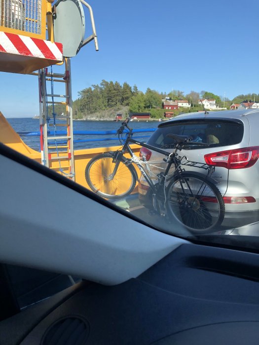 Cykel fastspänd på baksidan av en bil på en färja, med utsikt mot kusten.