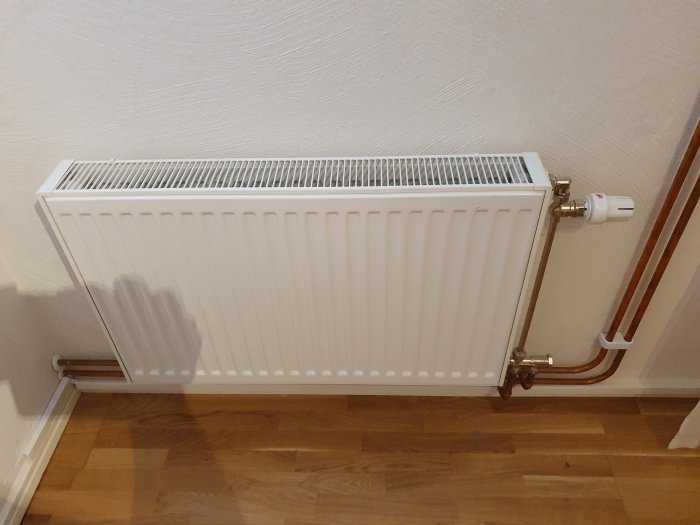 Vit vägghängd radiator med termostat och kopparledningar mot en ljus vägg och parkettgolv.
