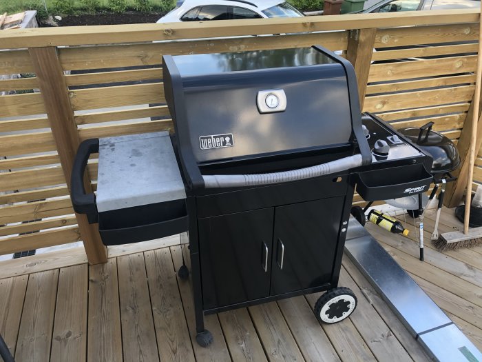 Begagnad svart Weber-grill på träaltan med grillverktyg och gasoltub.