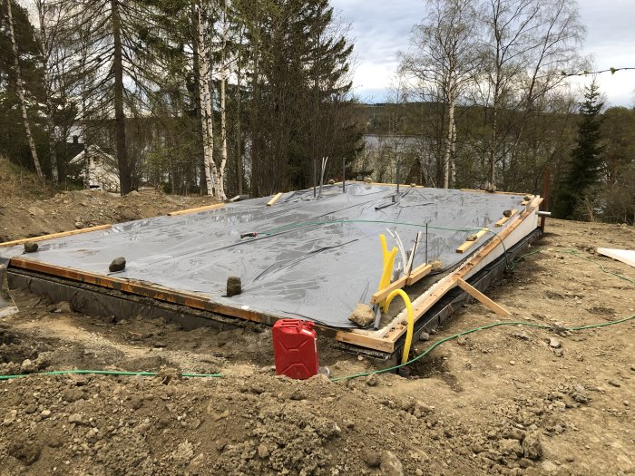 Nyggjuten betongplatta täckt med plast för källarnedgång, omgiven av byggmaterial och natur.