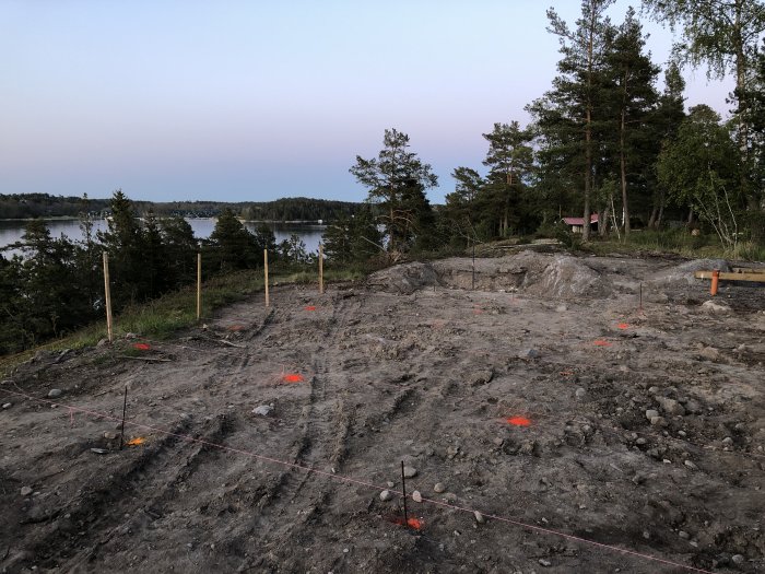 Uppmätt byggtomt med utplacerade träpinnar och snören samt utsikt över sjö och skog i skymningen.