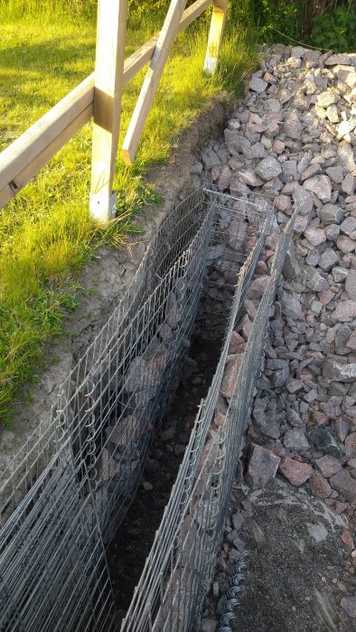 Gabionkorgar delvis fyllda med oregelbundna stenar bredvid en hög med finsprängt berg och gräsmatta.